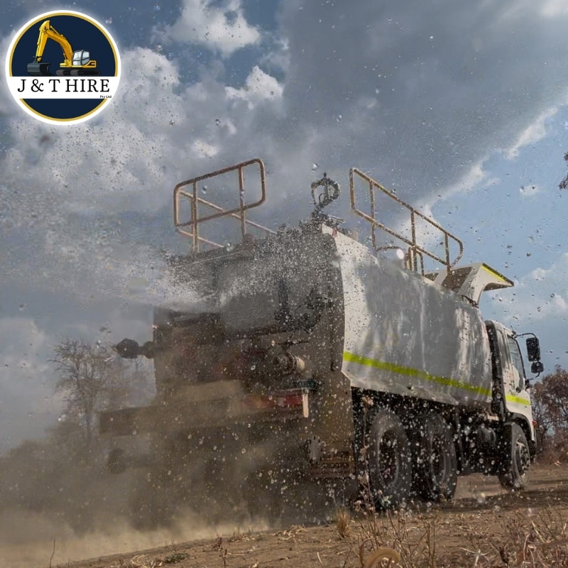 A water truck hire in Gunpowder sprays water over a dry landscape, creating a mist against a blue sky with scattered clouds, with the J&T Hire logo in the corner.